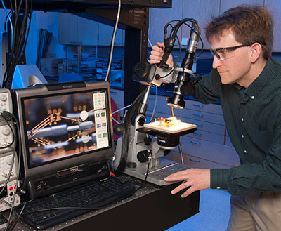 Dr. Ronald Polcawich performs high-resolution imaging and testing of a millimeter-scale robotic leg structure. The roughly 3-mm-long leg consists of segments of piezoelectric thin-film actuators and thin-film copper sections designed to mimic the kinetics of a leg with a "hip," "knee" and "ankle" joint sections. These "legs" have demonstrated the ability to move, lift (e.g., salt crystal in image), and resist impact into the silicon substrate. (Photo by Doug Lafon)